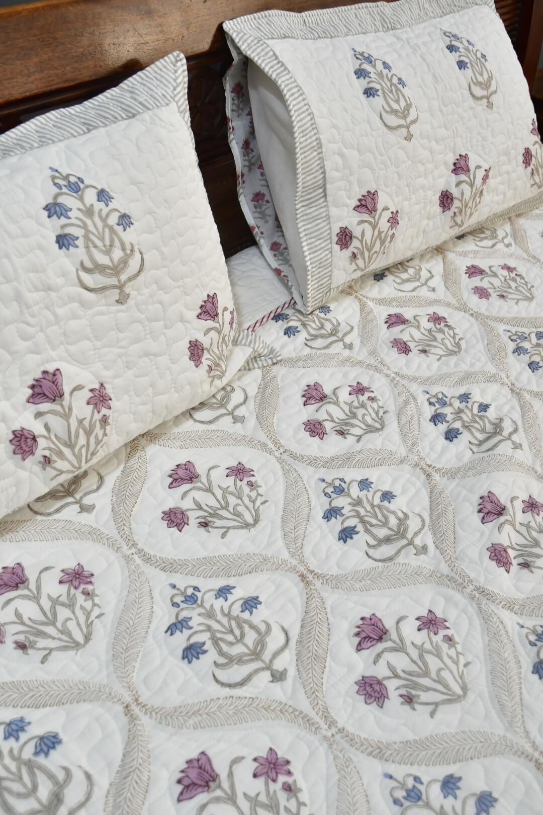 A quilted bedspread and matching pillow featuring a floral pattern in purple, blue, and beige on an off-white background, styled on a bed with a dark wood headboard.