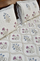 A quilted bedspread and matching pillow featuring a floral pattern in purple, blue, and beige on an off-white background, styled on a bed with a dark wood headboard.
