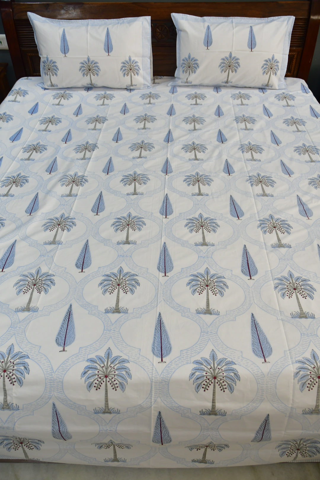 A bed dressed with a white quilted bedspread and matching pillowcases, both printed with a repeating pattern of blue palm trees and geometric medallions.