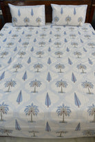 A bed dressed with a white quilted bedspread and matching pillowcases, both printed with a repeating pattern of blue palm trees and geometric medallions.