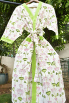 A white cotton wrap robe with a pink lotus and green lotus leaf pattern, featuring lime green trim and a matching tie belt, hanging on a wooden hanger outdoors.