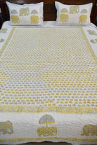 A quilted bedspread and matching pillowcases featuring a repeating block-print style elephant pattern in yellow and gray on a white background, styled on a bed with a dark wooden headboard.
