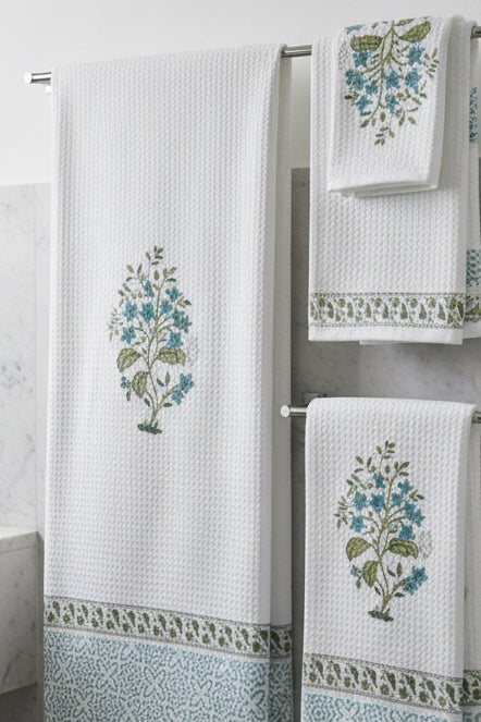 A white cotton tablecloth with blue and green floral block prints and a geometric border, shown alongside matching napkins on a dark blue surface.