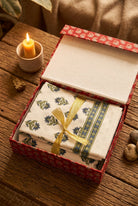 A folded white fabric textile with blue and green floral block-print style motifs and a blue striped border, tied with a gold ribbon, presented in an open red patterned gift box on a rustic wooden table with a lit candle nearby.