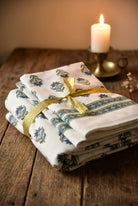 A stack of white cotton napkins with blue floral prints and a gold ribbon, tied on a rustic wooden table next to a lit candle.
