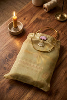 A folded light beige embroidered fabric pouch or small bag with a gold-tone handle and tassel detail, styled on a wooden tabletop beside a lit candle and spools of thread.