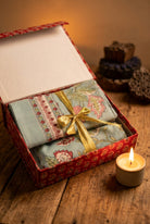 A folded patterned fabric item, likely a scarf or shawl, presented inside an open decorative gift box with a gold ribbon bow. The box sits on a rustic wooden table next to a lit candle, with warm, atmospheric background lighting.
