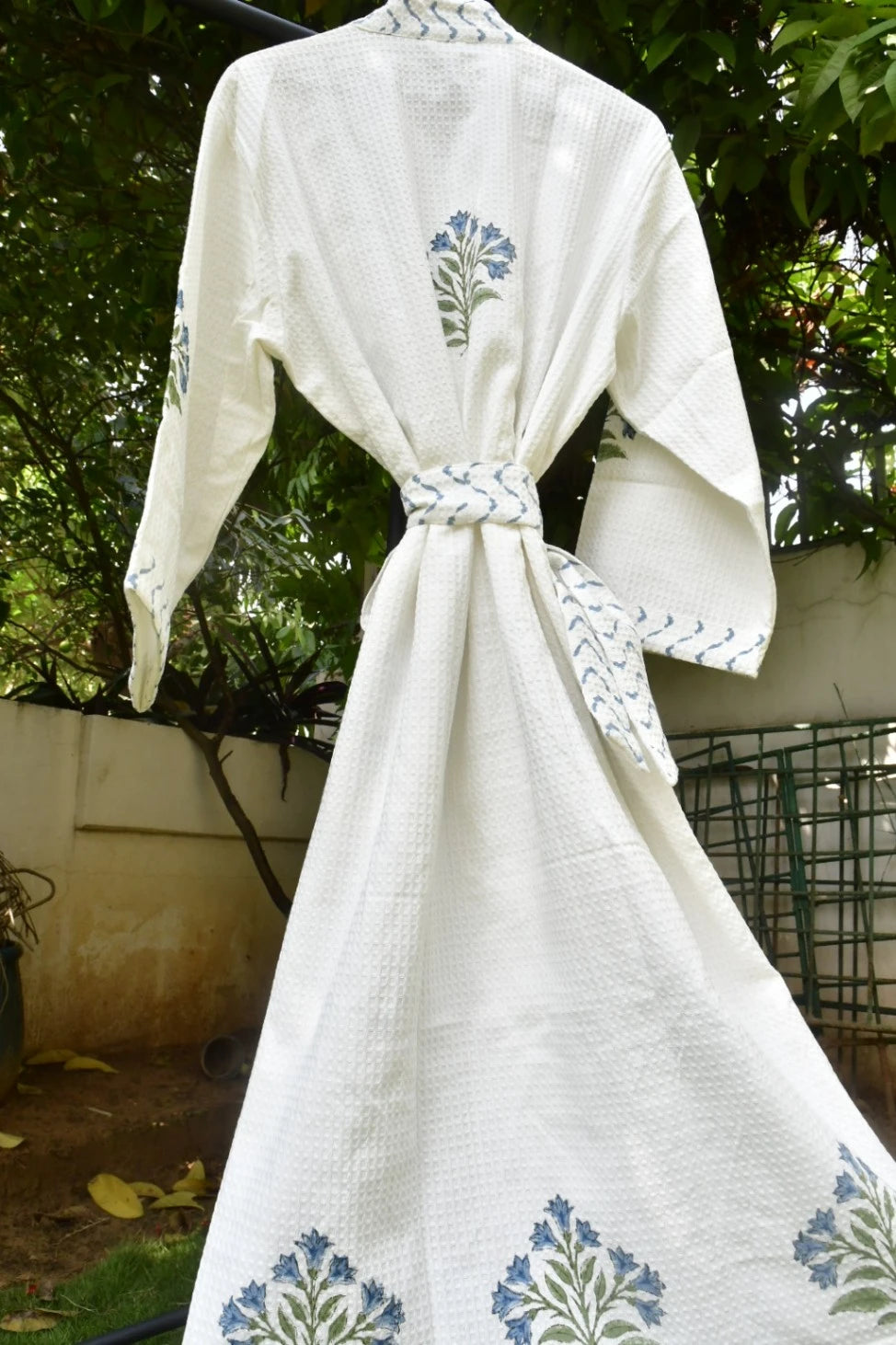 An off-white cotton kimono robe with blue and green floral block prints, tied at the waist with a matching printed belt, shown hanging outdoors against a background of green foliage.