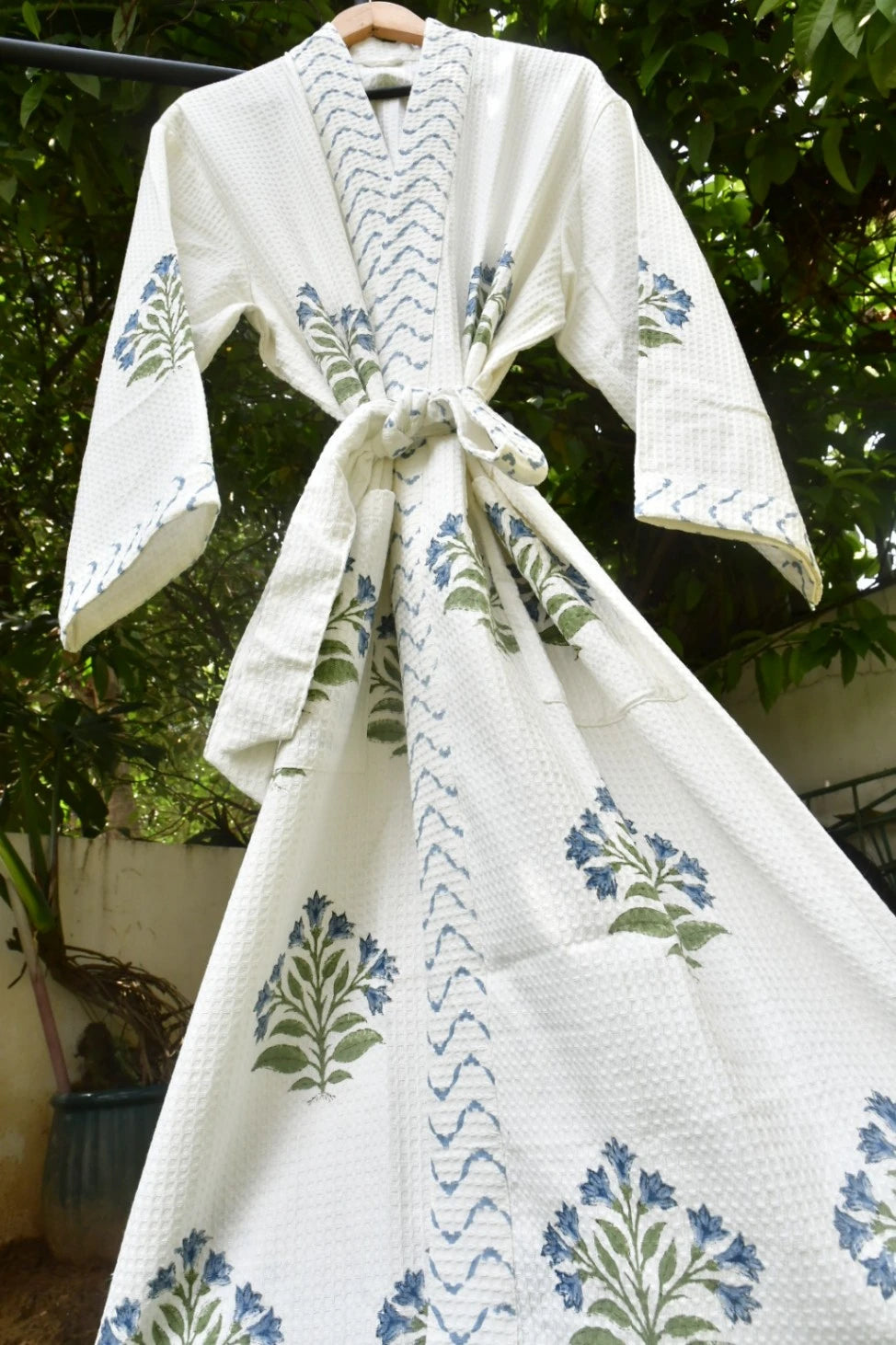 A white cotton robe with blue and green floral block prints and chevron patterns, hanging on a wooden hanger outdoors against a background of green foliage.