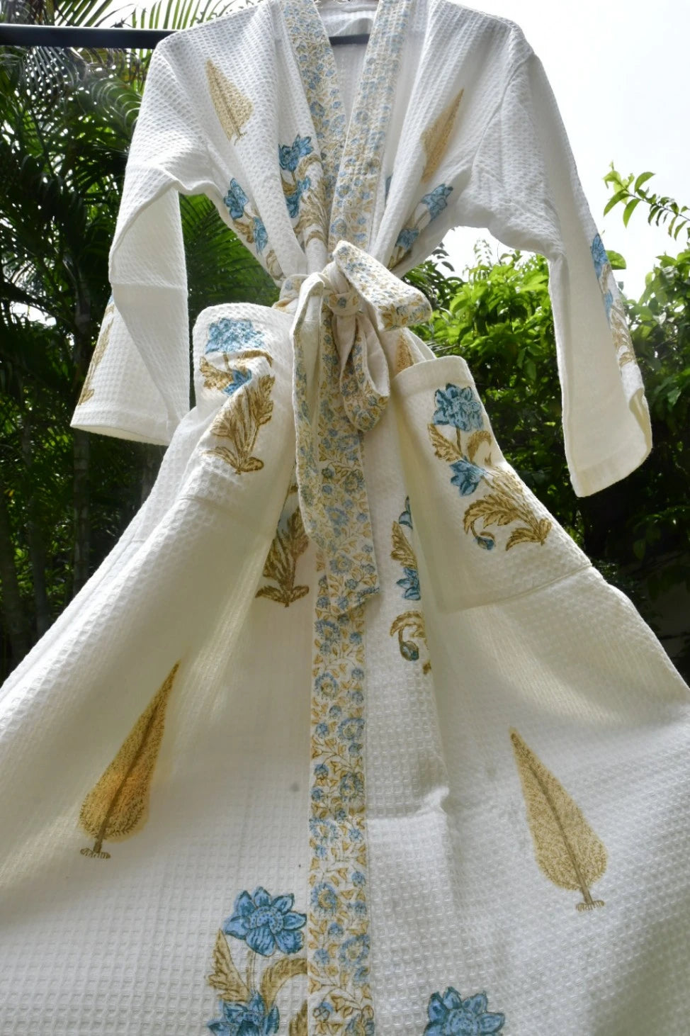 A white waffle-knit kimono robe with blue floral and gold leaf patterns, tied at the waist with a matching patterned belt, shown outdoors against a background of green foliage.