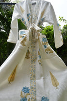 A white waffle-knit kimono robe with blue floral and gold leaf patterns, tied at the waist with a matching patterned belt, shown outdoors against a background of green foliage.