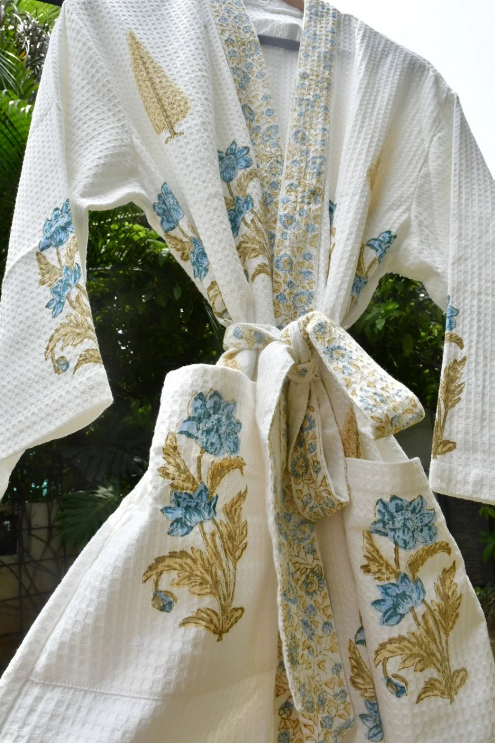A white waffle-knit kimono-style robe with blue and gold floral embroidery, tied at the waist, photographed outdoors against a blurred green background.