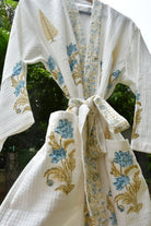 A white waffle-knit kimono-style robe with blue and gold floral embroidery, tied at the waist, photographed outdoors against a blurred green background.