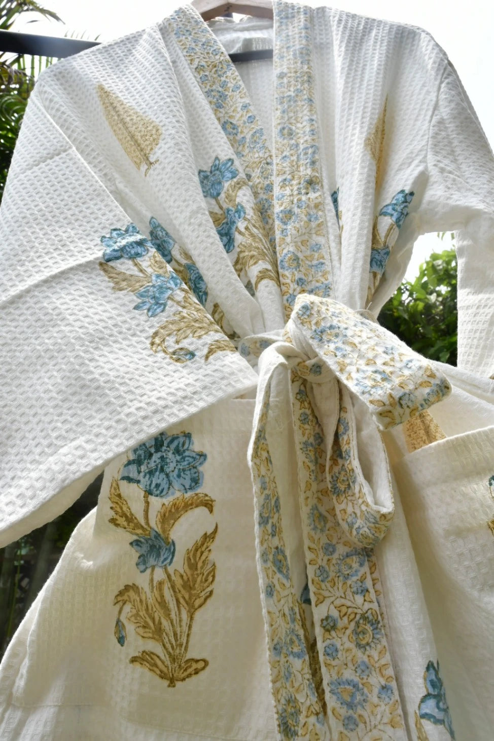 A white waffle-weave cotton robe with blue and gold floral embroidery, tied at the waist, shown hanging outdoors.