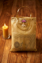 A beige organza and silk mixed fabric tote bag with a gold metallic handle, decorated with a small purple lotus flower and pearl-like beads, resting on a wooden table next to a lit candle.