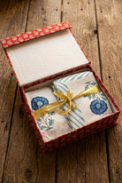 A gift box containing folded white textiles with blue floral and striped patterns, tied with a shiny gold ribbon, set against a dark background.