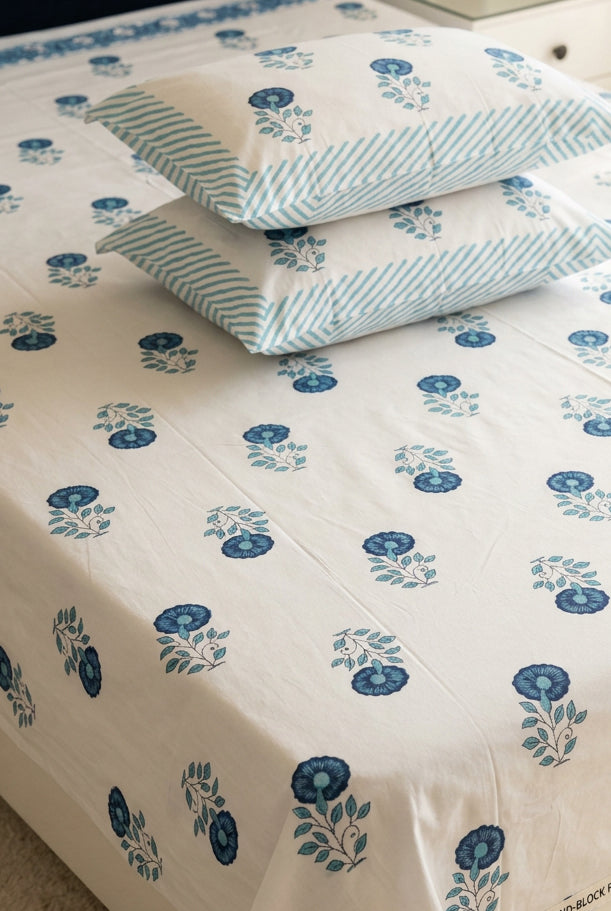 A bed made with a white bed sheet and matching pillowcases featuring a blue floral block print pattern. The sheet and two pillows have blue flowers with green leaves, while the other two pillows have blue and white stripes.