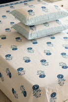 A bed made with a white bed sheet and matching pillowcases featuring a blue floral block print pattern. The sheet and two pillows have blue flowers with green leaves, while the other two pillows have blue and white stripes.