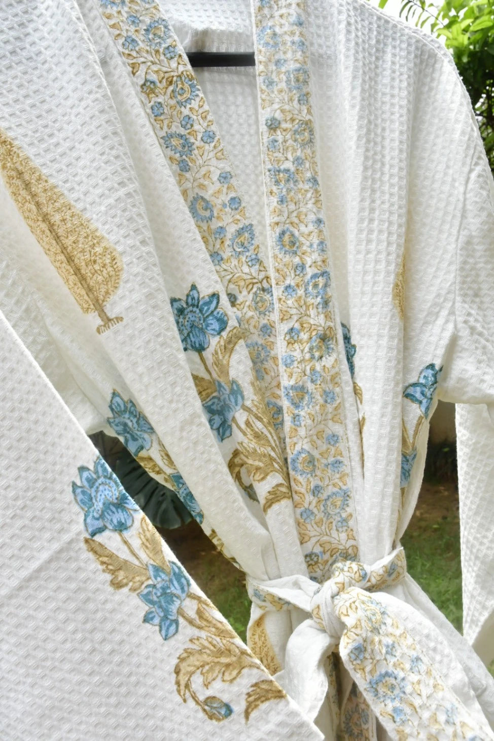 White waffle-weave robe with blue and gold floral embroidery, tied at the waist and shown outdoors with green foliage in the background.