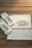 A set of three white cotton kitchen towels with blue and green floral embroidery and geometric patterns, laid out on a textured blue background.