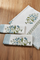 A set of white cotton napkins with blue floral prints, green leaves, and geometric borders, arranged on a textured blue background.