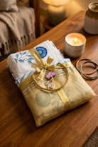 A folded yellow silk saree with a gold border and a pink lotus flower embellishment, tied with a gold ribbon and wrapped in a white cloth with blue floral patterns, displayed on a dark wooden surface.