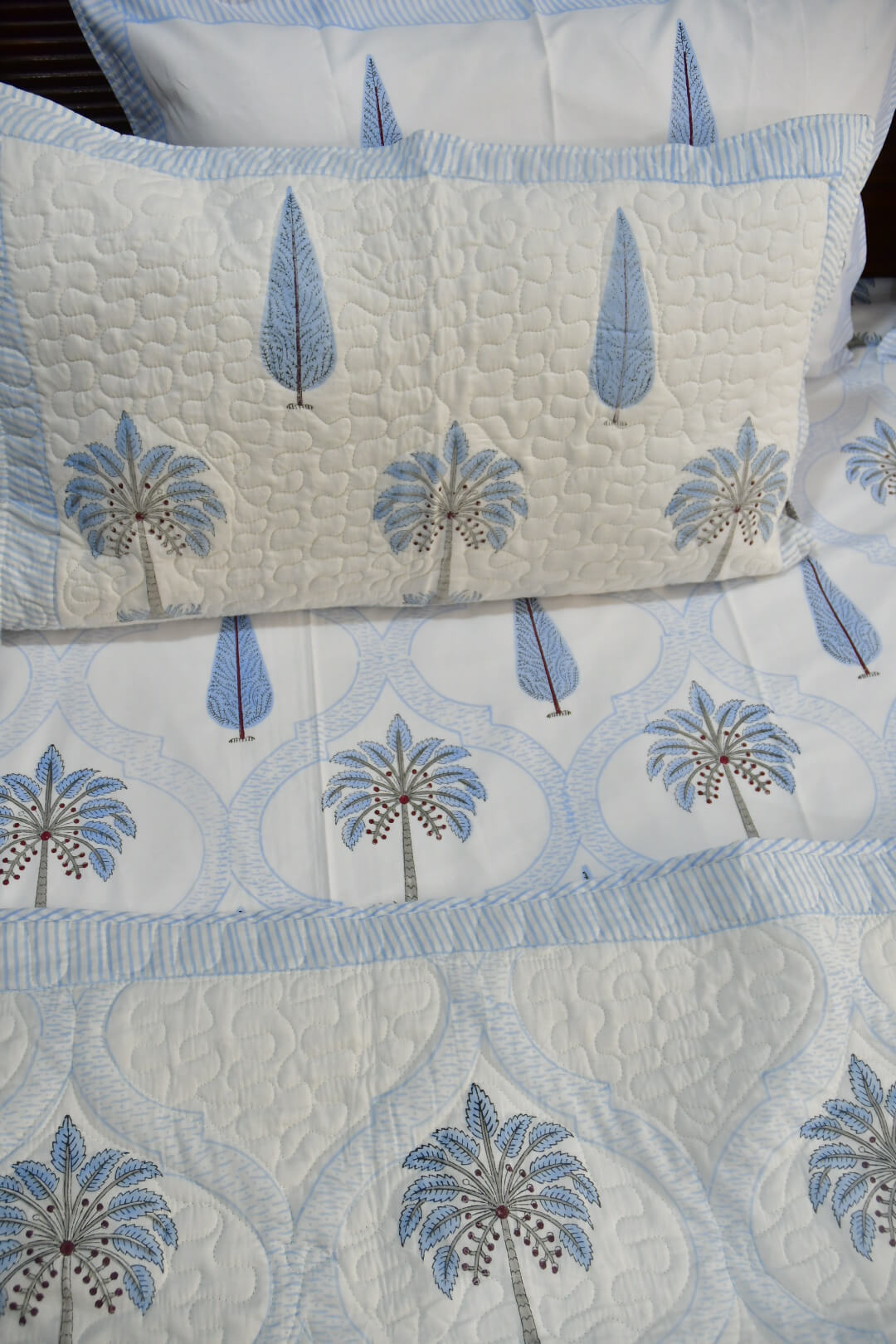 A close-up of a quilted bedding set featuring a cream-colored fabric with a light blue and gray palm tree pattern, complete with a matching pillowcase.