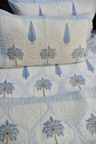 A close-up of a quilted bedding set featuring a cream-colored fabric with a light blue and gray palm tree pattern, complete with a matching pillowcase.