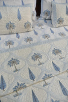 A quilted bedding set featuring a cream and light blue tropical pattern with palm trees and leaf motifs, styled with matching pillows.