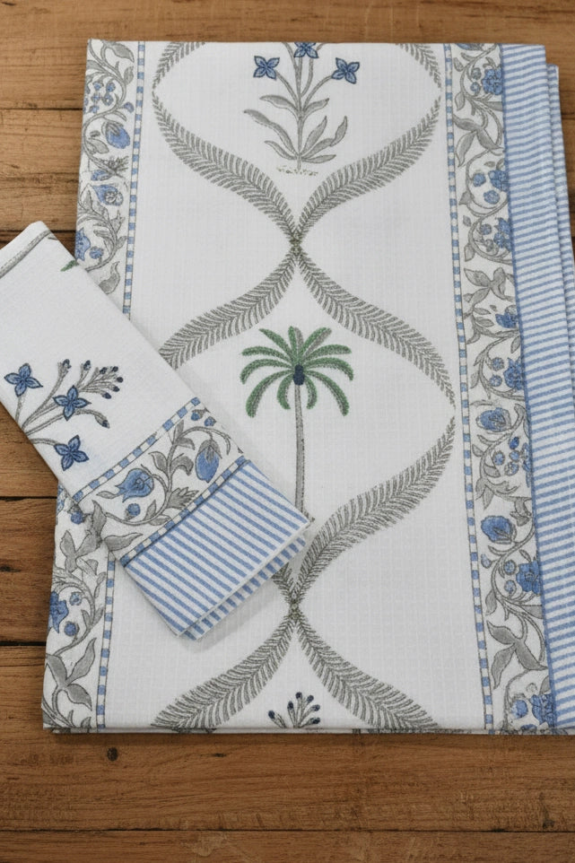 A folded white cotton fabric set, towel & napkin set, featuring blue floral prints, gray palm tree motifs, and blue-and-white striped borders, displayed on a rustic wooden surface.