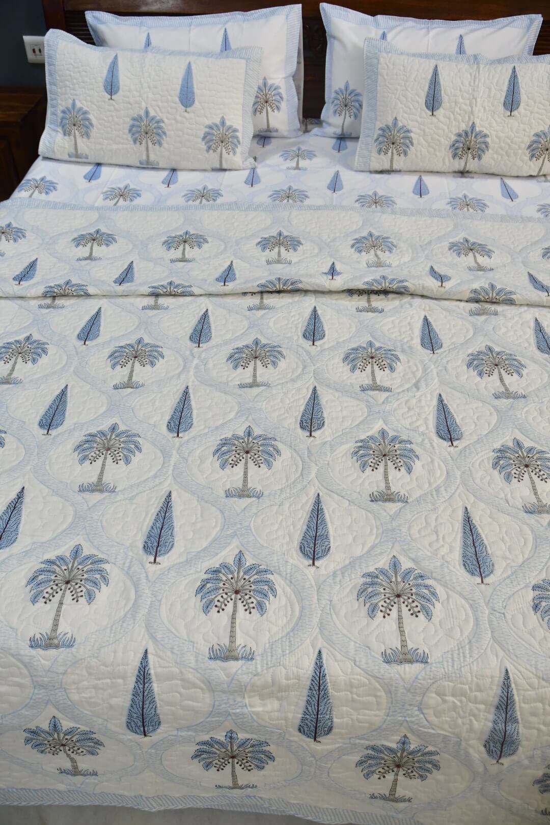 A quilted bedspread set featuring a repeating pattern of blue palm trees and teardrop motifs on a white background, styled on a bed with matching pillow shams.