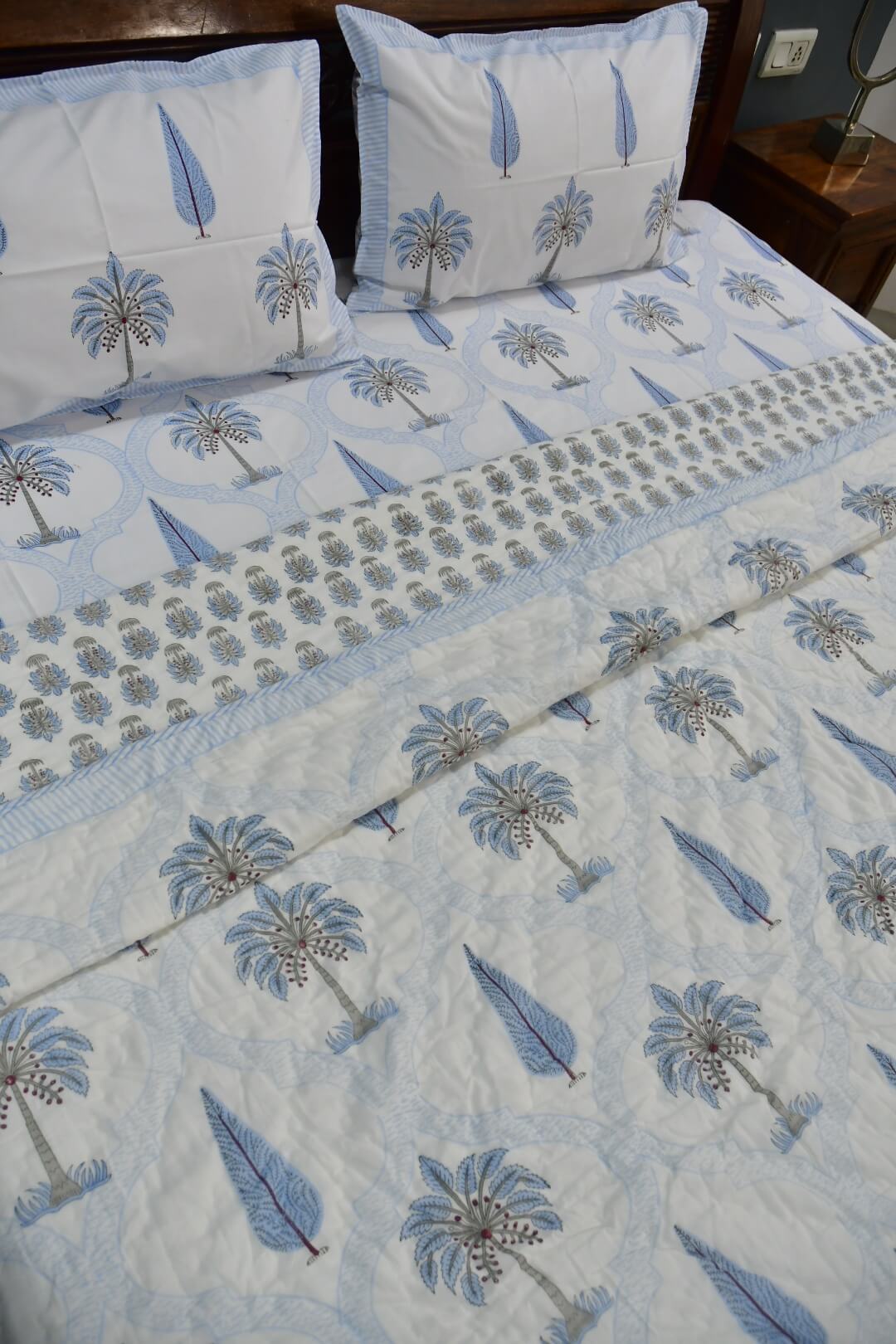 A light blue and white bedding set featuring a palm tree pattern, including a quilt, matching pillowcases, and a decorative runner, shown on a bed.