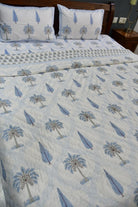 A bed dressed with a light blue and white quilted bedding set featuring a palm tree and feather pattern, with two matching pillowcases and a patterned runner, shown in a bedroom setting.