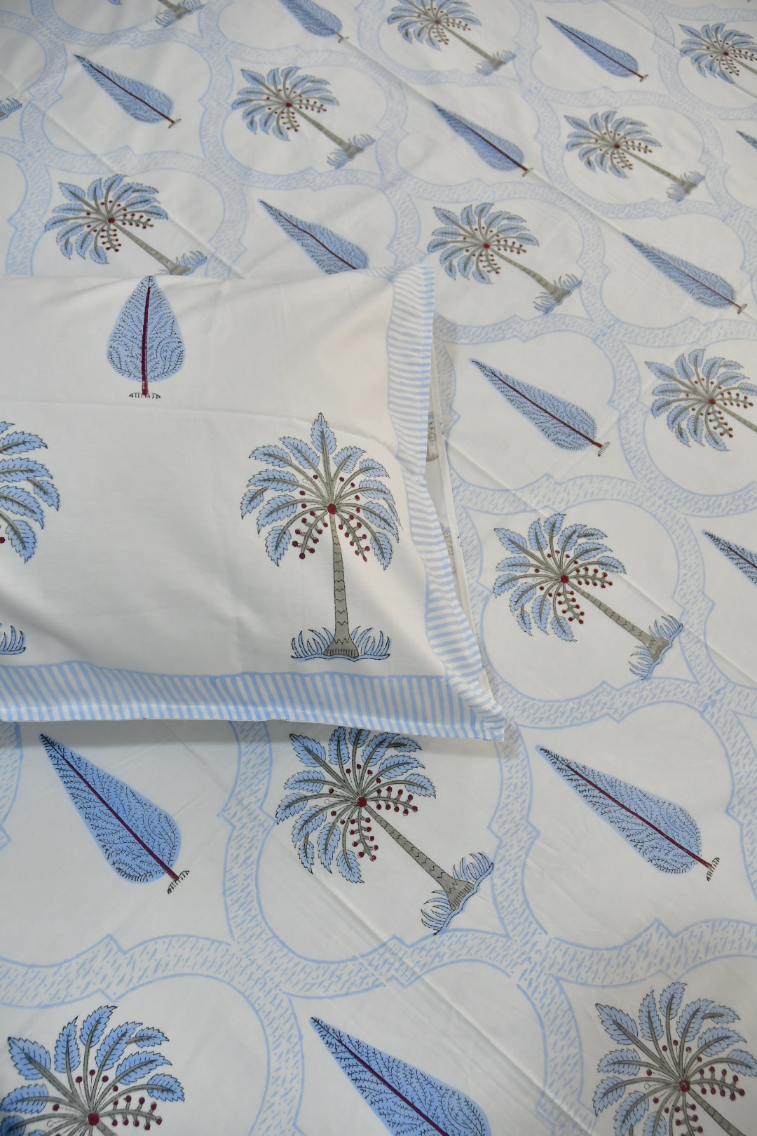 A close-up of a white quilted bedspread or comforter with a repeating palm tree and leaf pattern in light blue, medium blue, and gray, with a matching pillowcase partially visible.