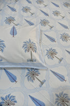 A close-up of a white quilted bedspread or comforter with a repeating palm tree and leaf pattern in light blue, medium blue, and gray, with a matching pillowcase partially visible.