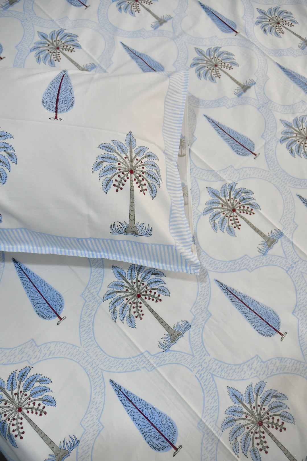 A close-up of a white quilted fabric with a repeating pattern of blue palm trees and leaf motifs, with a matching pillowcase partially visible.