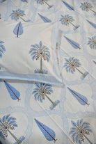 A close-up of a white quilted fabric with a repeating pattern of blue palm trees and leaf motifs, with a matching pillowcase partially visible.