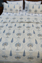 A quilted bedspread set featuring a repeating pattern of blue palm trees and teardrop motifs on a white background, styled on a bed with matching pillow shams.