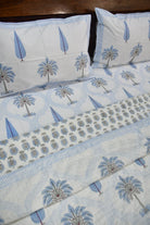 A close-up of a bed dressed in a white quilted bedding set with a light blue and gray palm tree block print, including two matching pillowcases and a folded comforter or duvet.
