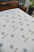 A white dohar featuring a repeating pattern of stylized blue and beige lotus flowers, shown on a bed in a bedroom setting.