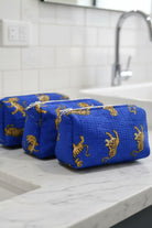 Three blue zippered cosmetic pouches with a gold tiger print, arranged diagonally on a blue textured blanket in a bedroom setting.