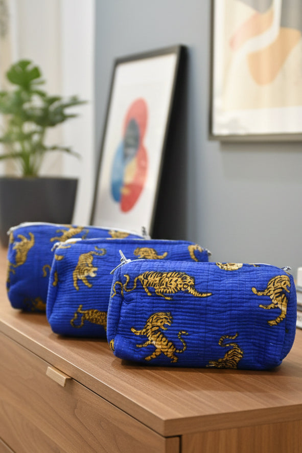 Three blue zippered cosmetic pouches with a gold tiger print, arranged on a blue textured blanket in a bedroom setting.
