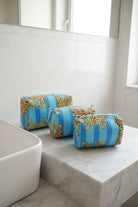 Three striped cosmetic pouches with a blue and gold pattern are arranged on a white marble bathroom counter next to a sink.