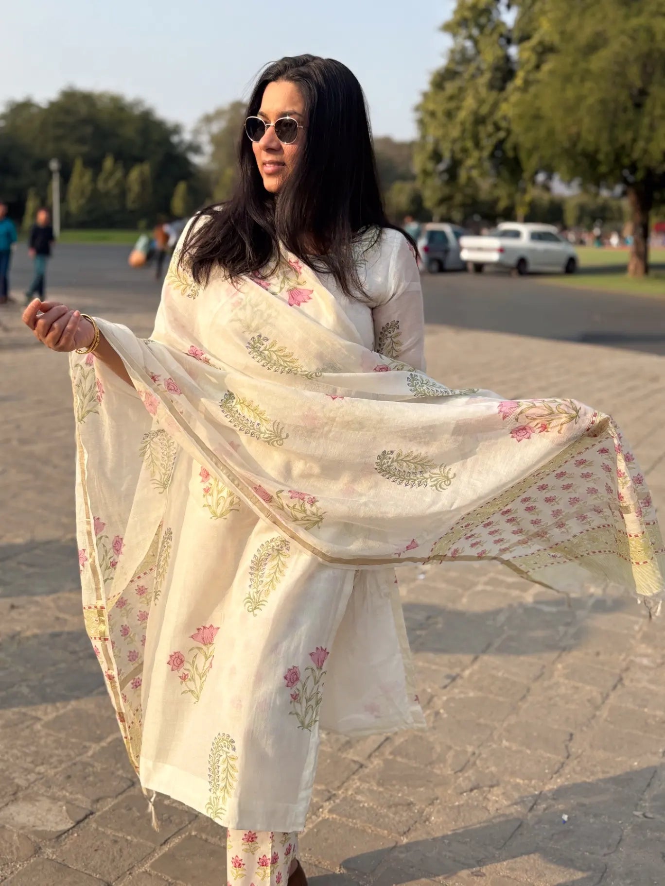 A woman outdoors wearing a light cream/off-white traditional outfit with pink floral and green leaf prints, holding out a matching printed dupatta. The background shows a paved walkway, trees, and distant cars.