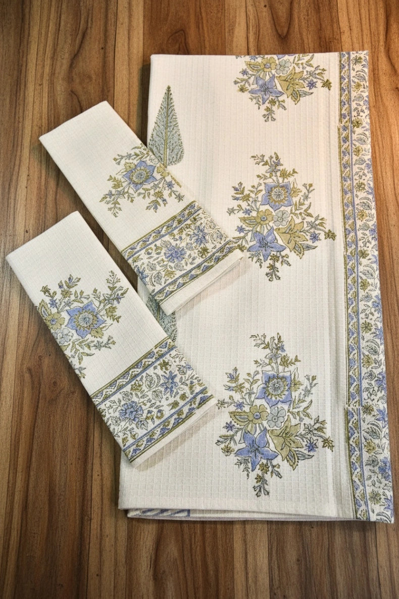 A set of white cotton table linens, including a napkin and two checkered placemats, featuring blue and green floral motifs and borders, displayed on a wooden surface.