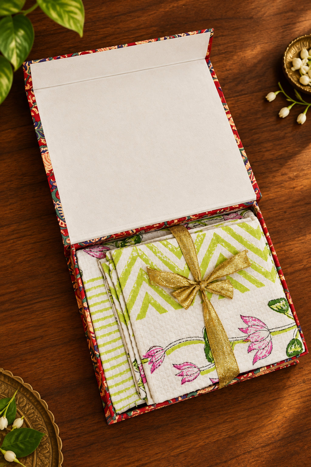 A gift box containing folded patterned napkins or small towels, tied with a gold ribbon. The visible fabric has green chevron stripes and pink floral motifs on a white background, set on a dark wooden surface.