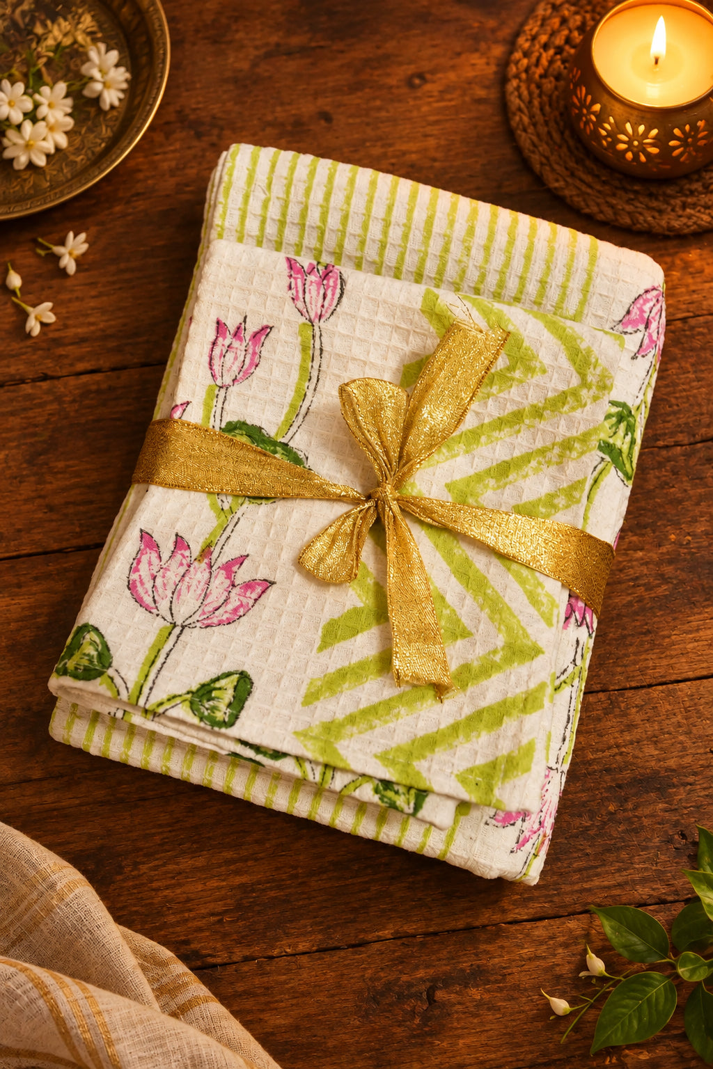 A folded white cotton kitchen towel or napkin featuring green and white stripes, pink tulips, and green leaves, tied with a shiny gold ribbon bow, displayed on a dark wooden surface.