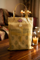 A yellow-gold sheer fabric handbag with a metallic gold handle, decorated with a small pink flower and white pearl-like beads, resting on a dark wooden surface.