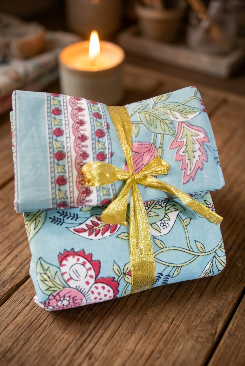 A folded quilted fabric piece with a light blue background and a multicolor floral and bird print, tied with a shiny gold ribbon, displayed on a rustic wooden surface with a blurred lit candle in the background.