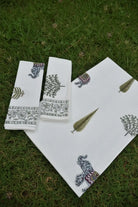 A set of white cotton towels featuring hand-block printed elephant and leaf motifs in muted green, gray, and purple, laid out on a green grass background.